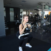 Focused and determined, a woman performs a kettlebell squat with A90 Hip Bands in a well-equipped gym, showcasing strength and dedication in her fitness routine.